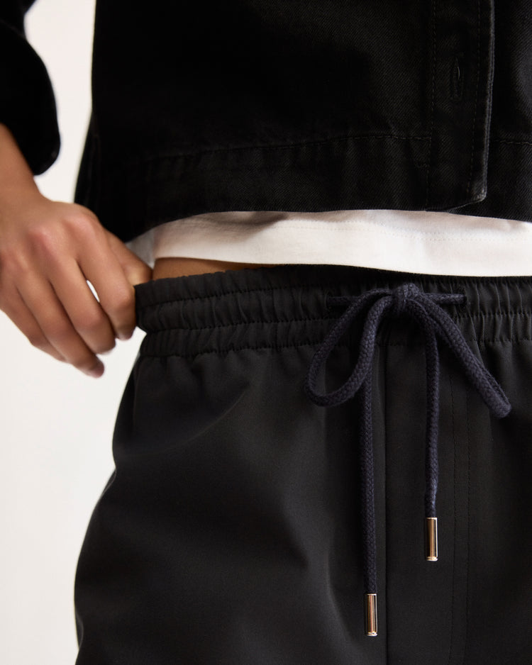 A person stands with one hand on their hip, wearing a white shirt, a tailored black jacket, and Arlo Mott's Morgan Long Shorts in Black. The focus is on the waist area and the tied drawstrings of the shorts.