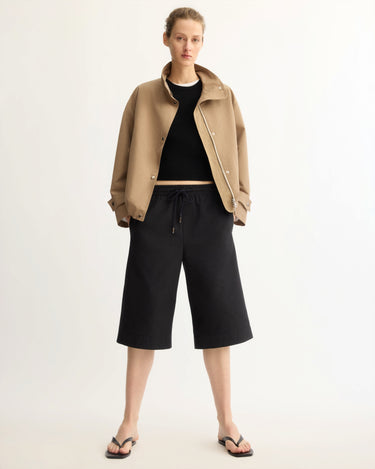 A person faces forward in a tailored tan jacket, black crop top, Arlo Mott's Morgan Long Shorts in Black, and black sandals against a plain light background.