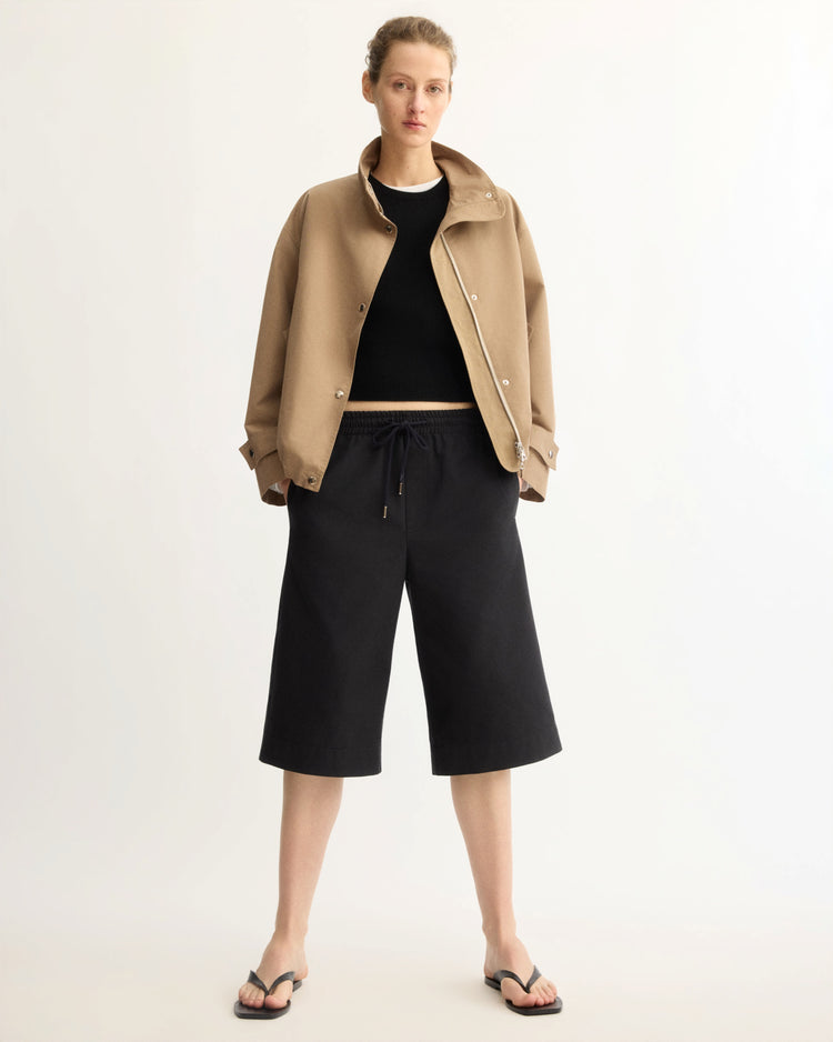 A person faces forward in a tailored tan jacket, black crop top, Arlo Mott's Morgan Long Shorts in Black, and black sandals against a plain light background.