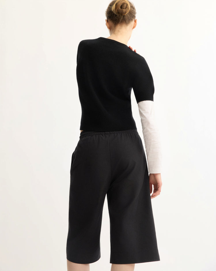 A person with their hair in a bun stands against a plain white background, wearing Arlo Mott’s Morgan Long Shorts in Black, paired with a black short-sleeve sweater layered over a long-sleeve white shirt.