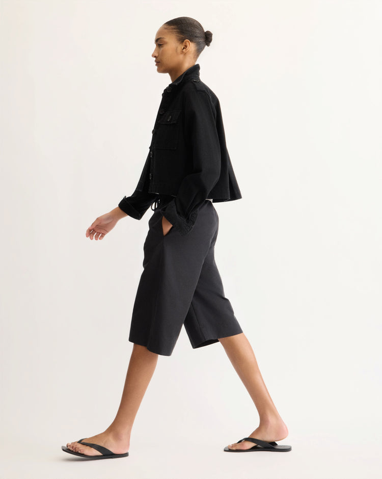 A person with hair in a bun wears the Arlo Mott Henry Short Shirt Jacket in Black, paired with knee-length black shorts and black flip-flops, walking in profile against a plain light background.