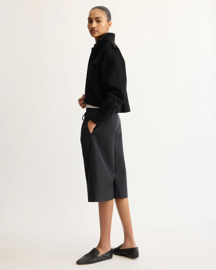Against a plain background, a woman in profile wears the Arlo Mott Henry Short Shirt Jacket in Black with knee-length black culottes and flat shoes, her hair tied back and hands in her pockets—highlighting the jacket’s ideal layering style.