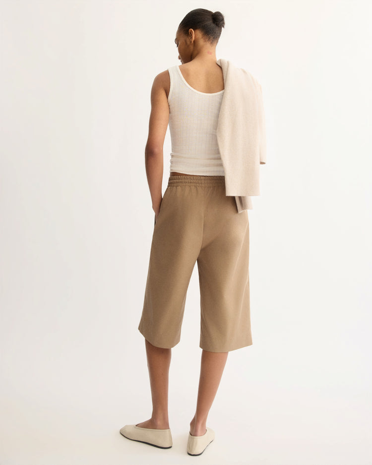 A person faces away, wearing a white sleeveless top, Arlo Mott’s Morgan Long Shorts in Khaki, and beige slip-on shoes. A light beige sweater is draped over their shoulder, and their neat hair is styled in a bun.