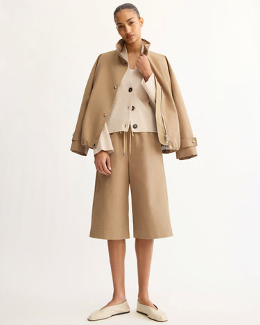 A woman stands against a plain background, wearing a tan Italian gabardine jacket, a beige buttoned cardigan, Arlo Mott's Morgan Long Shorts in Khaki, and cream-colored slip-on shoes.