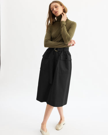 A woman stands against a plain white background, wearing an olive green turtleneck and the Arlo Mott Felix Utility Midi Skirt in Black with utility pockets and a belted waist. She pairs it with cream flats, arms crossed and head slightly tilted.