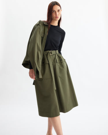 A woman wears a black long-sleeve top and the Arlo Mott Felix Utility Midi Skirt in Olive, featuring utility pockets, with a matching green jacket over one shoulder, posing against a plain white background.