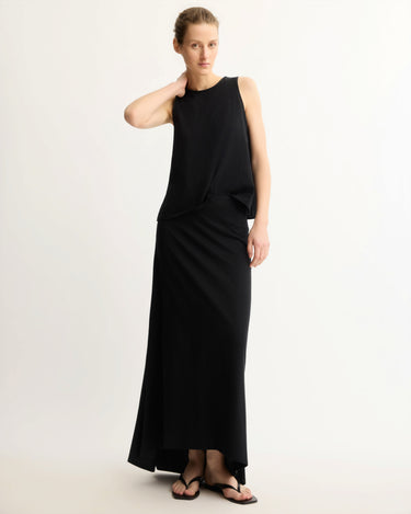 Against a plain light background, a person with light skin and blonde hair pulled back wears a sleeveless black cotton jersey top, black sandals, and the Arlo Mott Beekman Maxi Skirt in Black with a subtle mermaid hem. One hand touches their neck.