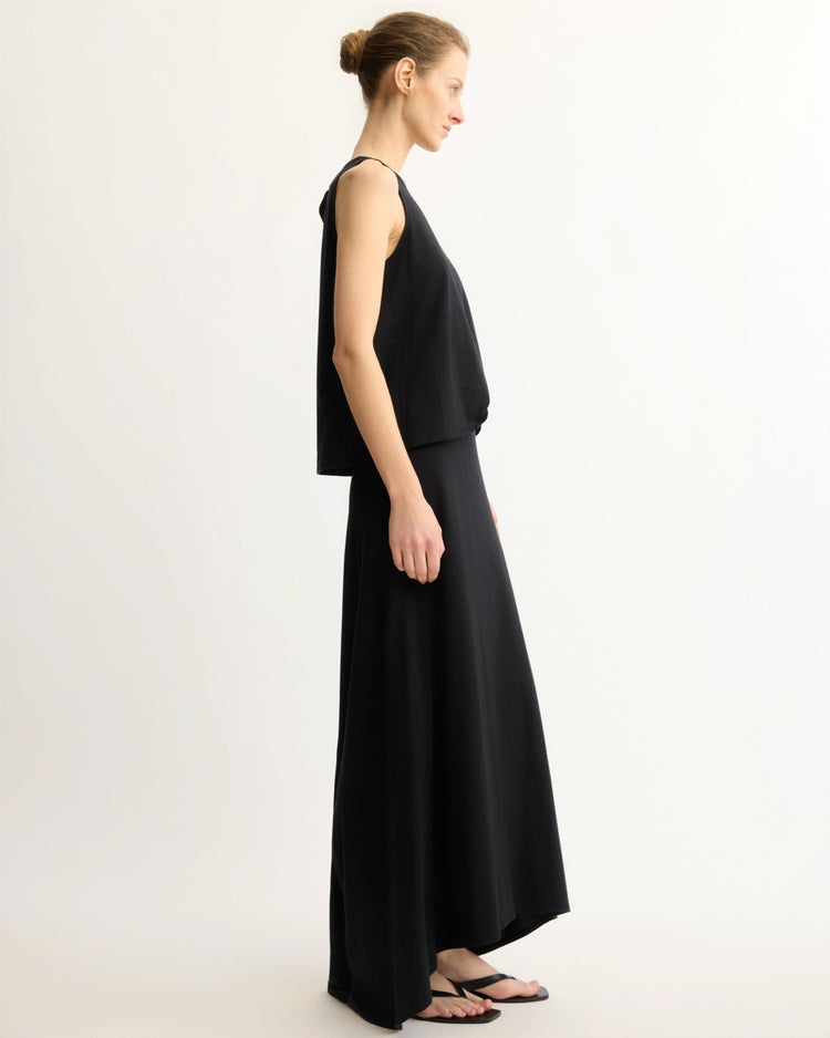 A woman with blonde hair in a bun stands in profile against a plain background, wearing a loose black sleeveless top, black sandals, and the Arlo Mott Beekman Maxi Skirt in Black.