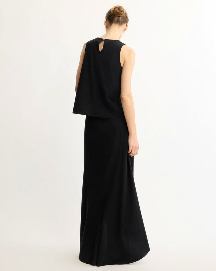 A woman with light skin and blond hair in a neat bun is seen from behind wearing the Arlo Mott Beekman Maxi Skirt in Black and a sleeveless black cotton jersey top, standing against a plain white background.