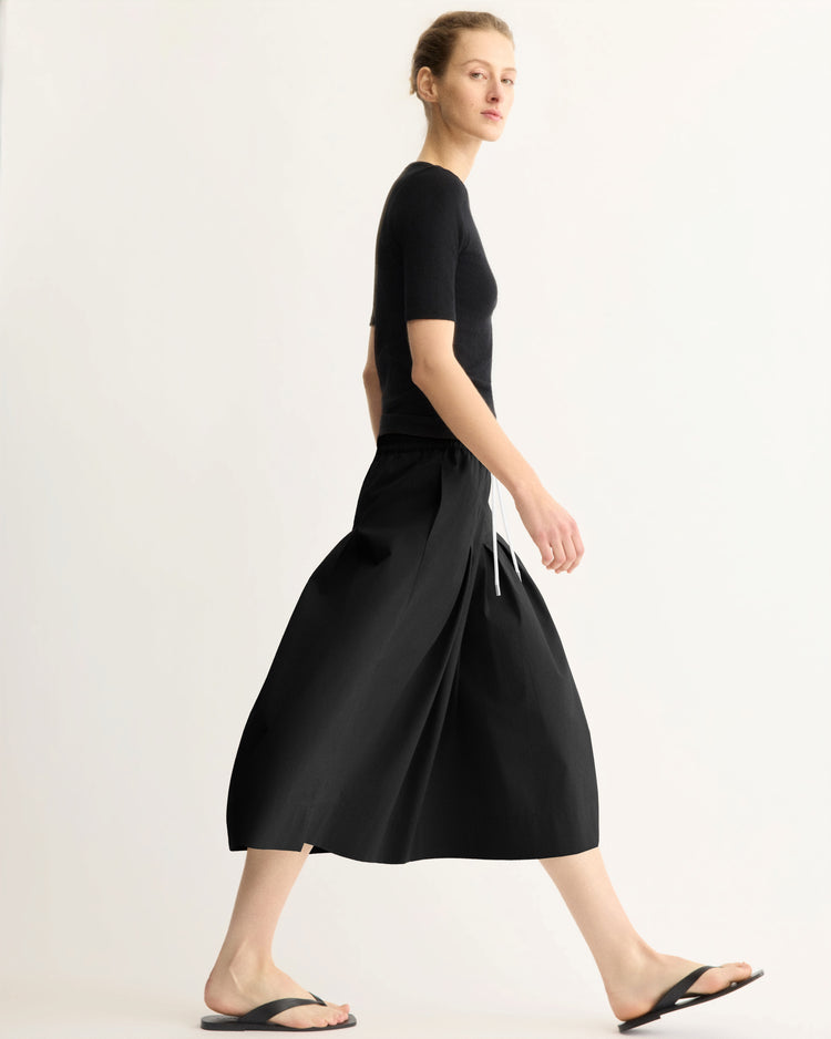 A woman in a black short-sleeve top, Arlo Mott Gates Full Midi Skirt in Black with a white drawstring waist, and black flip-flops walks against a plain, light background.