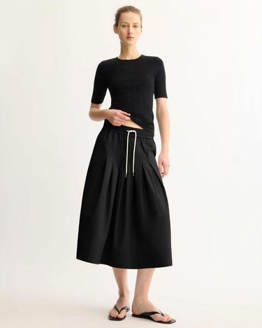 A woman with her hair tied back and a neutral expression wears a fitted black short-sleeve top, Arlo Mott's Gates Full Midi Skirt in Black, and black sandals against a plain light background.
