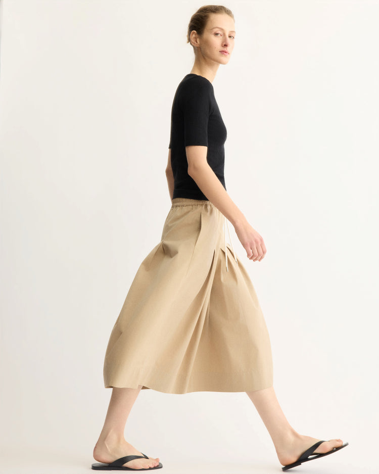 A woman in profile wears a fitted black short-sleeve top, Arlo Mott’s Gates Full Midi Skirt in Khaki with a drawstring waist, and black flip-flops against a plain light background.