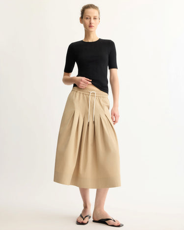 A person stands against a plain background wearing a fitted black short-sleeve top, Arlo Mott’s Gates Full Midi Skirt in Khaki with pleats and a drawstring waist, and black flip-flop sandals.