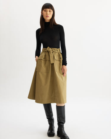 A woman faces forward in a black long-sleeve turtleneck, the Arlo Mott Felix Utility Midi Skirt in Khaki with utility pockets and waist tie, and tall black boots, all set against a plain white background.
