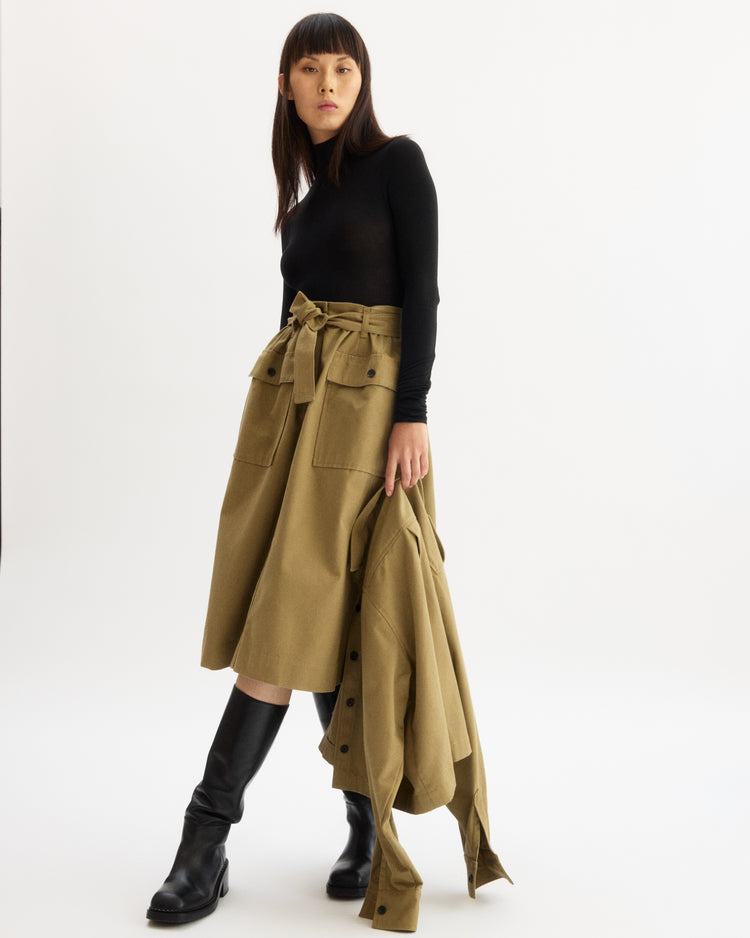 A woman stands against a white background wearing the Arlo Mott Chambers Long Sleeve Body Suit in Black, paired with a high-waisted khaki midi skirt with large pockets and a tie belt, knee-high black boots, and holding a matching khaki coat.