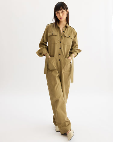 A woman stands in a studio on a white background, wearing the Arlo Mott Walker Utility Jumpsuit in Khaki, featuring front pockets and buttons. She has her hands in her pockets and looks toward the camera.