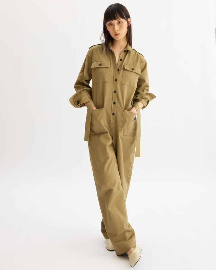A woman stands in a studio on a white background, wearing the Arlo Mott Walker Utility Jumpsuit in Khaki, featuring front pockets and buttons. She has her hands in her pockets and looks toward the camera.