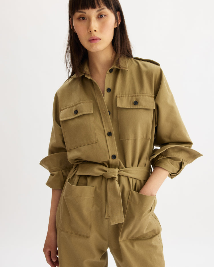 A person wears the Arlo Mott Walker Utility Jumpsuit in Khaki, featuring long sleeves, black buttons, chest pockets, and a tied belt, with one hand in their pocket against a plain white background.