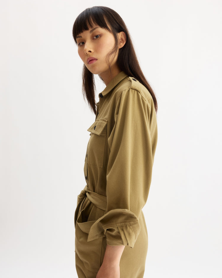 A woman with straight dark hair and bangs stands in profile against a white background, confidently wearing the Arlo Mott Walker Utility Jumpsuit in Khaki.