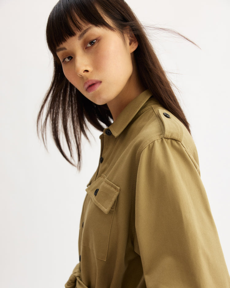 A woman with long dark hair and bangs poses confidently in the Arlo Mott Walker Utility Jumpsuit in Khaki against a plain light background, looking directly at the camera.