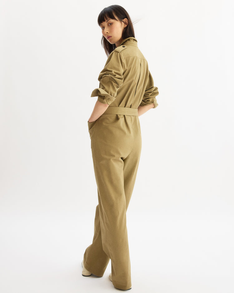 A woman with long dark hair stands slightly turned, hands in the pockets of her khaki Walker Utility Jumpsuit by Arlo Mott, sleeves rolled up, looking over her shoulder against a plain white background. Made in New York.