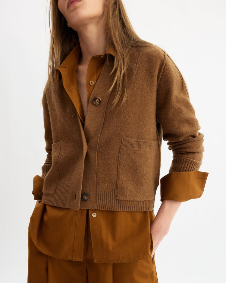 A person wears the Arlo Mott Perry Boxy Cardigan in Cinnamon over a brown button-up shirt with rolled sleeves, standing against a plain white background. Their face is partially out of frame.