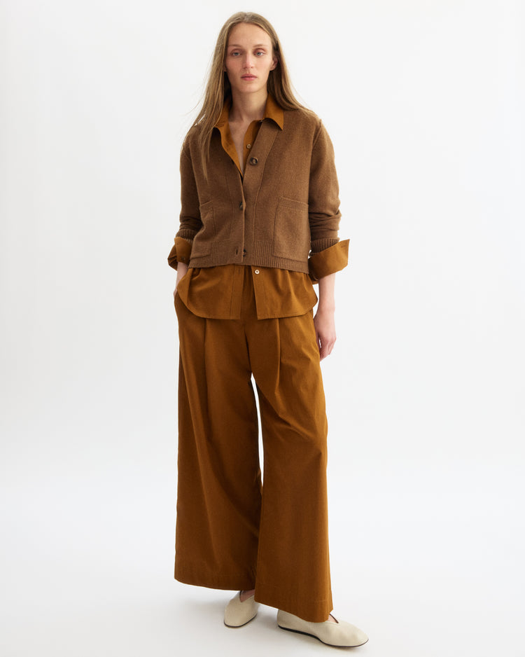 A woman with long straight hair and a neutral expression stands against a white background wearing Arlo Mott’s Perry Boxy Cardigan in Cinnamon, a matching button-up shirt, wide-legged brown pants, and off-white shoes.