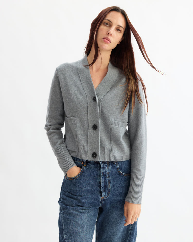 A woman with long brown hair wears the Arlo Mott Perry Boxy Cardigan in Heather Grey and blue jeans, standing against a plain white background with her hands in her pockets.