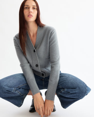 A woman with long brown hair squats in the Arlo Mott Perry Boxy Cardigan in Heather Grey and blue wide-leg jeans, gazing neutrally into the camera against a plain white background.