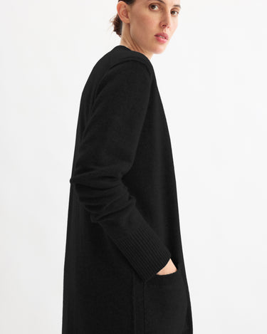 A person models the Arlo Mott Sidney Duster Cardigan in Black, standing sideways with one hand in a pocket and looking over their shoulder against a plain white background.