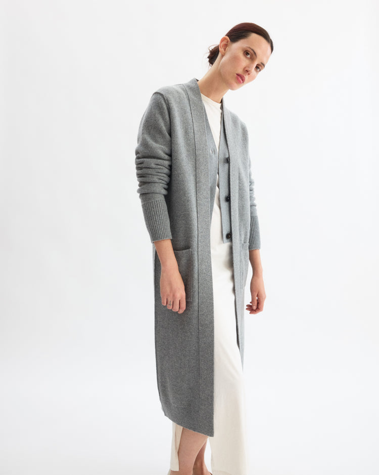 A woman wears the Arlo Mott Sidney Duster Cardigan in Heather Grey, a long wool-cashmere blend, over a white dress. She stands against a plain white background, gazing slightly to the side with a neutral expression.