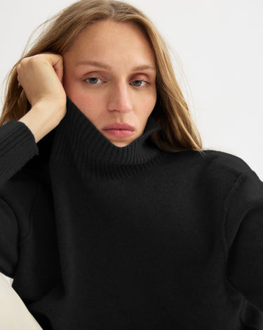 A person with long, light brown hair wears the Monroe Boxy Turtleneck in Black by Arlo Mott, resting their head on one hand and looking thoughtful against a plain light background.