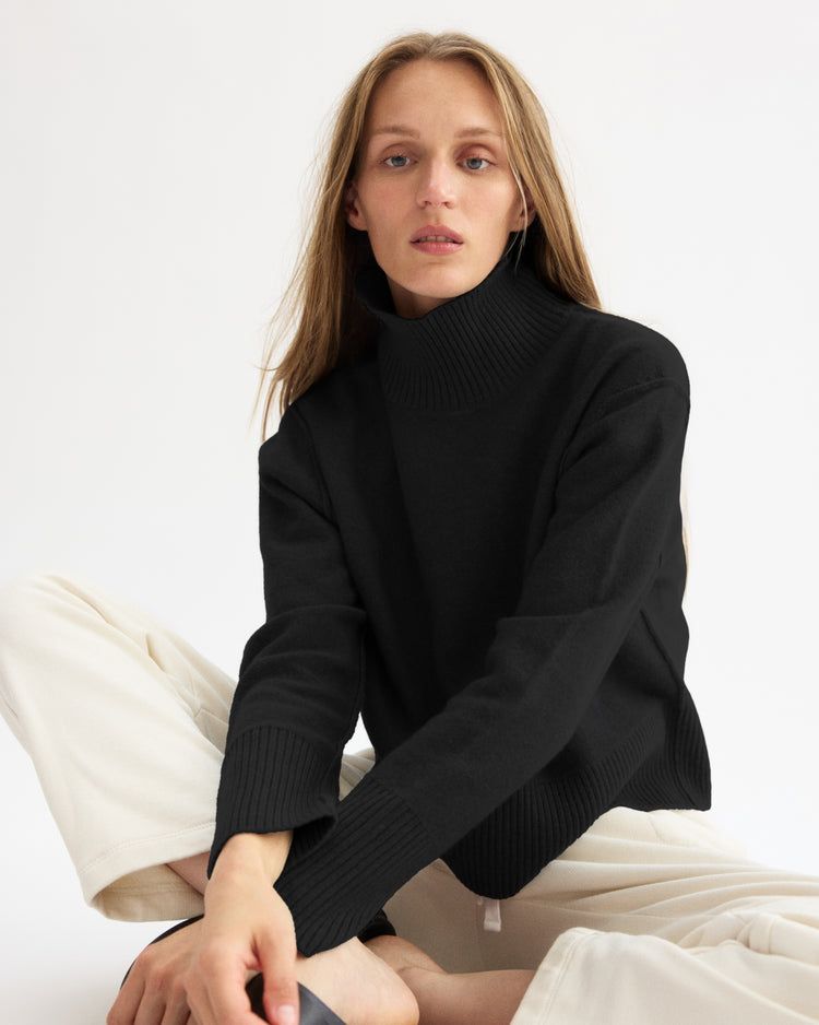 A person with long, light brown hair sits on the floor against a plain white background, wearing the Arlo Mott Monroe Boxy Turtleneck in Black and cream-colored pants, looking at the camera with a neutral expression.
