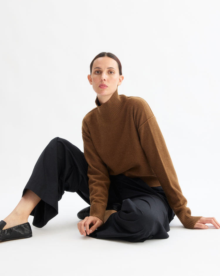A woman with dark hair in a low bun sits on the floor against a white background, wearing the Arlo Mott Monroe Boxy Turtleneck in Cinnamon, loose black pants, and black flats, looking directly at the camera with a neutral expression.