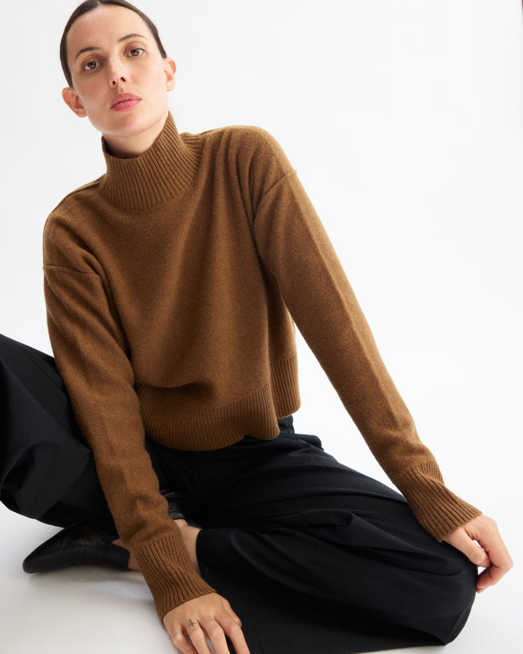 A person with short hair, wearing the Arlo Mott Monroe Boxy Turtleneck in Cinnamon and black pants, sits on the floor with one arm resting on their knee against a plain white background.