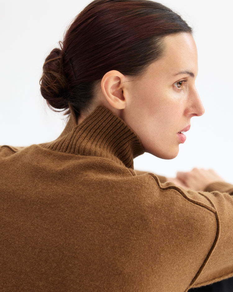 A woman with dark hair in a low bun wears the Arlo Mott Monroe Boxy Turtleneck in Cinnamon, shown in profile against a plain light background.