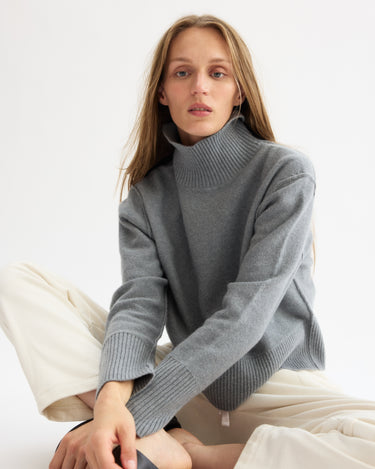 A woman with long, light brown hair wears the Arlo Mott Monroe Boxy Turtleneck in Heather Grey and cream pants, sitting on the floor against a plain white background and looking at the camera with a neutral expression.