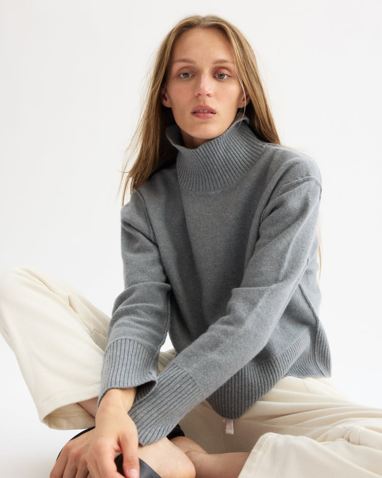 A woman with long, light brown hair wears the Arlo Mott Monroe Boxy Turtleneck in Heather Grey and cream pants, sitting on the floor against a plain white background and looking at the camera with a neutral expression.