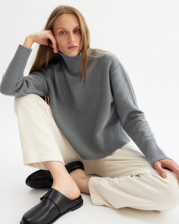 A person with long light brown hair sits on the floor against a white background, wearing the Arlo Mott Monroe Boxy Turtleneck in Heather Grey, cream pants, and black slip-on shoes, looking at the camera with a neutral expression.