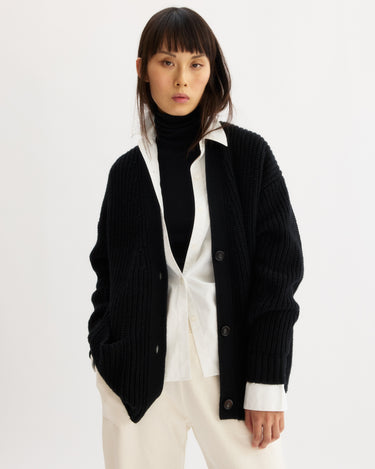 A woman with straight brown hair models the Arlo Mott Bedford Grandpa Cardigan in Black, layered over a white button-up and black wool turtleneck. She stands with one hand in her pocket against a plain white background.