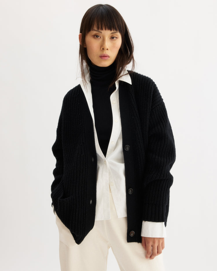 A woman with straight brown hair models the Arlo Mott Bedford Grandpa Cardigan in Black, layered over a white button-up and black wool turtleneck. She stands with one hand in her pocket against a plain white background.