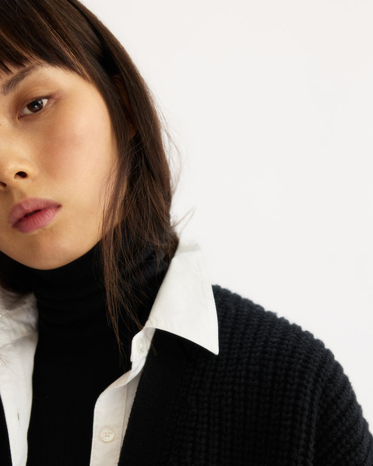 A close-up photo of a person with straight dark hair in a white collared shirt layered under the Arlo Mott Bedford Grandpa Cardigan in Black, gazing slightly away with a neutral expression against a plain light background.
