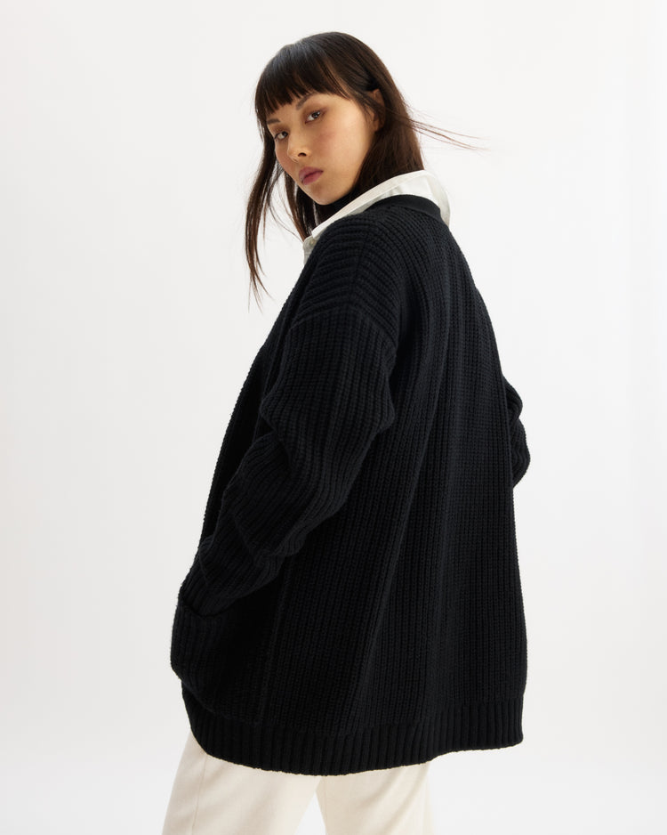A woman with straight dark hair models the Arlo Mott Bedford Grandpa Cardigan in Black over a white shirt and light pants, standing slightly turned against a plain white background and looking over her shoulder at the camera.