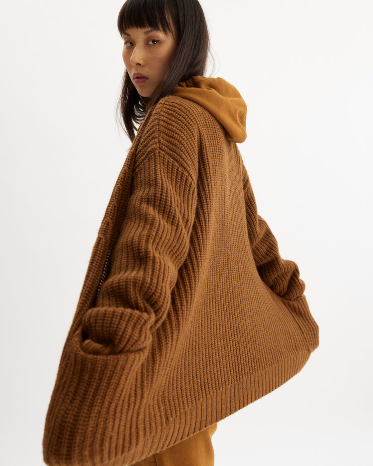 A woman with straight dark hair and bangs wears the Arlo Mott Bedford Grandpa Cardigan in Cinnamon, an oversized textured brown wool sweater with wide sleeves and a hood, standing against a plain white background.