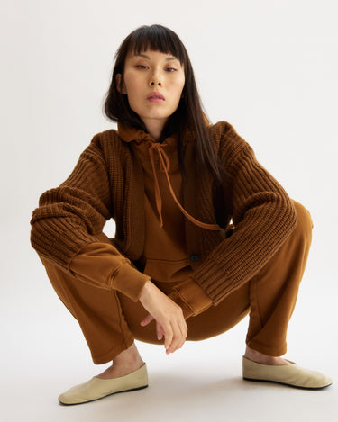 A woman with long dark hair squats confidently in Arlo Mott's Bedford Grandpa Cardigan in Cinnamon, styled over matching layers and beige slip-on shoes against a plain white background.