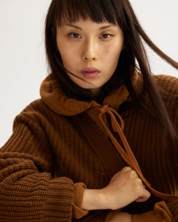 A woman with straight dark hair and bangs wears the Arlo Mott Bedford Grandpa Cardigan in Cinnamon, holding its drawstring and looking intently at the camera against a plain light background.