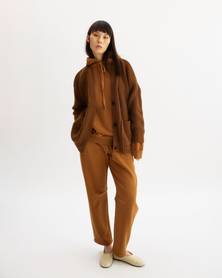 A woman with straight dark hair and bangs poses in Arlo Mott’s Bedford Grandpa Cardigan in Cinnamon over a brown hoodie and sweatpants, paired with beige slippers, against a plain white background.