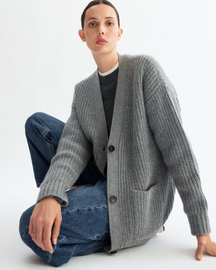 A person with short hair sits on the floor against a plain white background, wearing Arlo Mott’s Bedford Grandpa Cardigan in Heather Grey over a dark shirt and blue jeans.