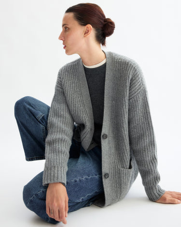 A person with dark hair tied back wears the Arlo Mott Bedford Grandpa Cardigan in Heather Grey, paired with a dark gray shirt and blue jeans, sitting on the floor with one knee up against a plain white background.