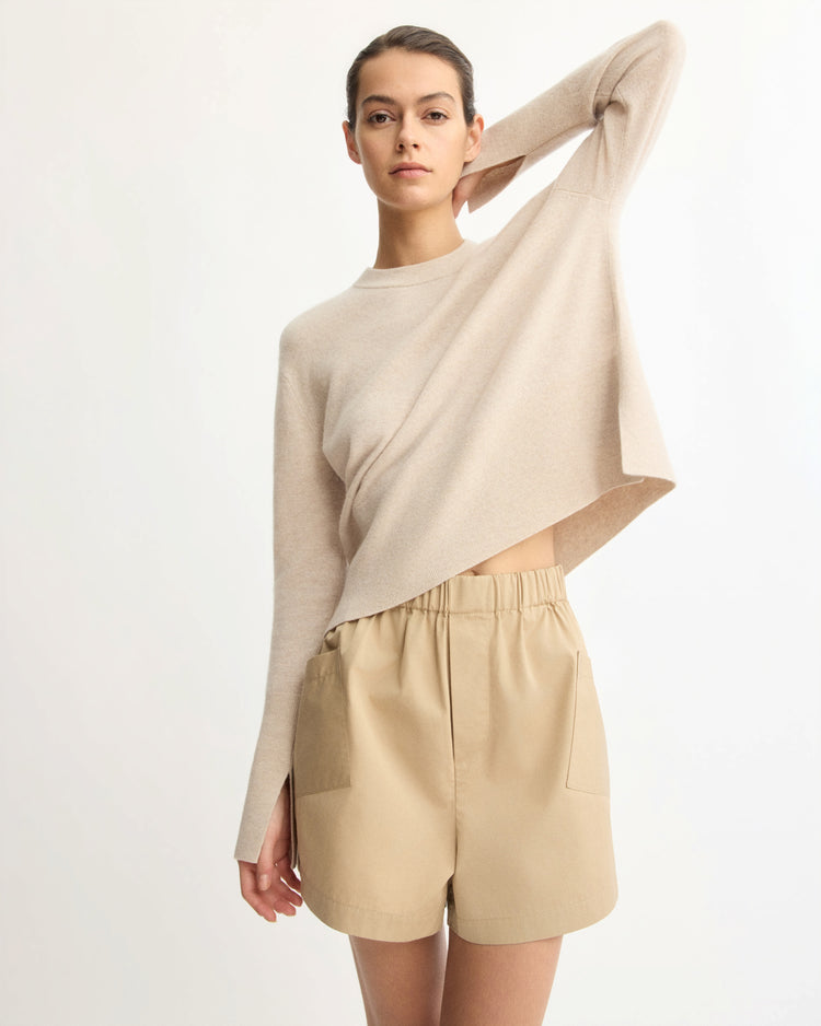 A woman poses confidently against a plain background in the Arlo Mott Warren Classic Crew Neck in Beige and tan high-waisted shorts with pockets, with one arm raised to touch the back of her head.
